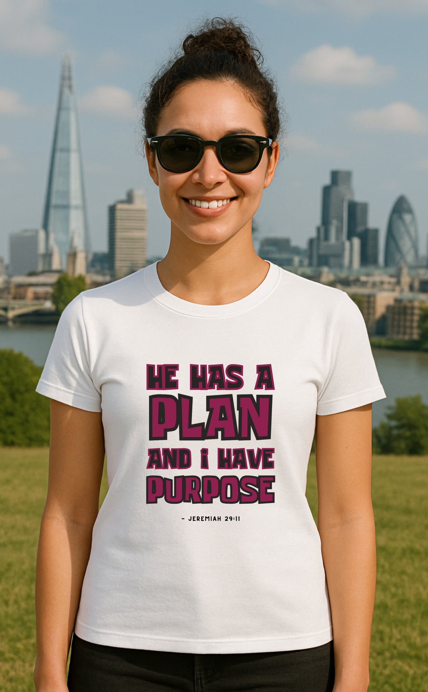 Smiling woman wearing white 'He has a plan and I have a purpose - Jeremiah 29:11' t-shirt from VerseWear by L’authority with bold typography – 'He has a plan and I have a purpose" and a smaller typology of - 'Jeremiah 29:11' – featuring black and deep prink, burgundy like colours with London skyline in the background