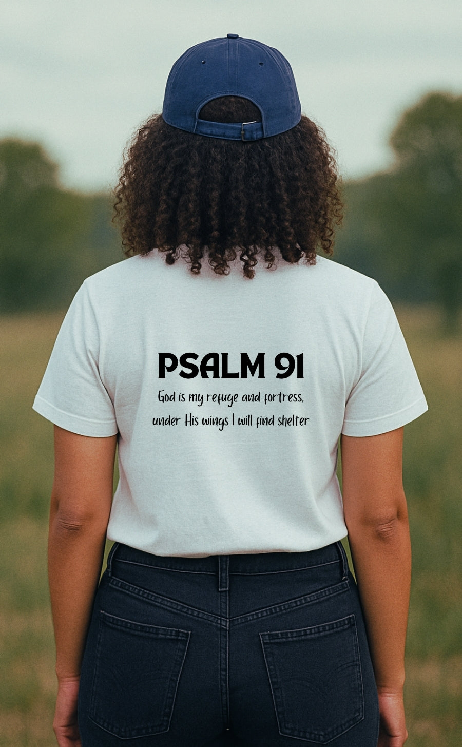 A young woman wearing a white "Refuge Code" Christian t-shirt from VerseWear by L'authority, seen from the back in a peaceful countryside setting. The shirt displays the Bible verse “Psalm 91 – God is my refuge and fortress, under His wings I will find shelter,” blending faith and style in a clean, minimalist design.