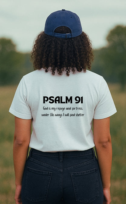 A young woman wearing a white "Refuge Code" Christian t-shirt from VerseWear by L'authority, seen from the back in a peaceful countryside setting. The shirt displays the Bible verse “Psalm 91 – God is my refuge and fortress, under His wings I will find shelter,” blending faith and style in a clean, minimalist design.