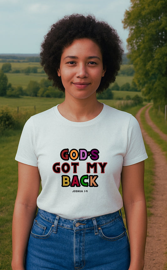 A young woman wearing a white “God’s Got My Back” t-shirt by VerseWear by L'authority, featuring bold multicoloured text and the Bible verse Joshua 1:9. She stands confidently on a countryside path with green fields in the background, embodying strength, faith, and modern Christian streetwear.
