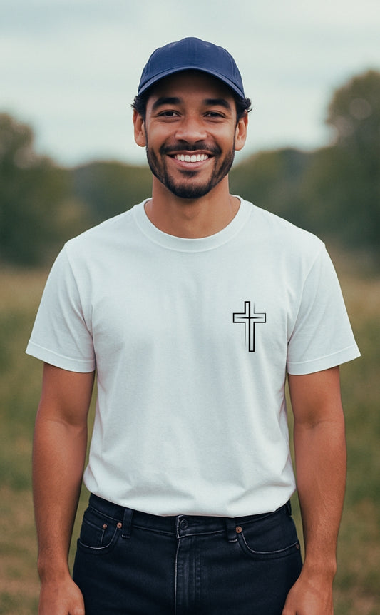 A smiling young Black man wearing a white Christian t-shirt from VerseWear by L’authority with a minimalist black cross symbol on the left chest. He stands outdoors in a serene, grassy countryside setting under soft natural light, radiating confidence and faith.
