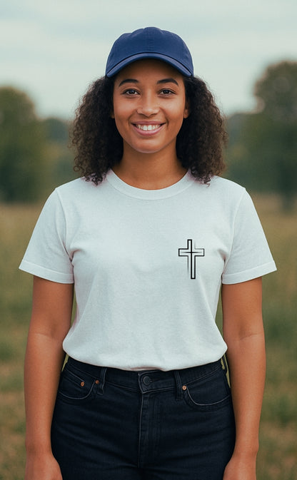 A young woman wearing a white Christian t-shirt from VerseWear by L'authority, featuring a minimalist black cross symbol on the left chest. She stands confidently in a peaceful rural setting, smiling softly—blending faith, simplicity, and modern streetwear style.